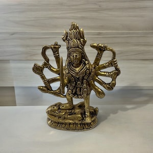 May include: A gold-colored metal statue of a Hindu deity with multiple arms holding various objects. The deity is standing on a base with a decorative design.