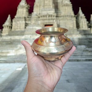 May include: A gold-colored metal pot with intricate designs sits in a hand, with a large, ornate, white building in the background.
