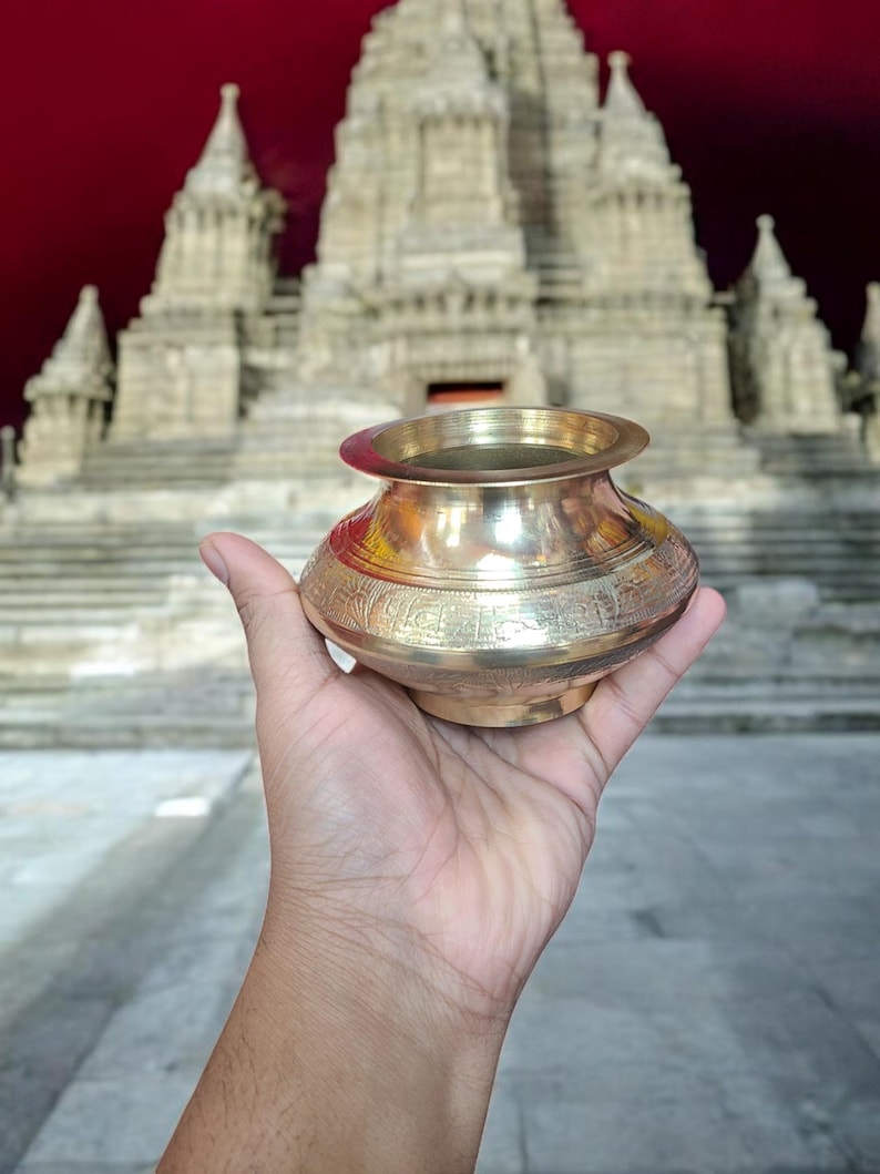 May include: A gold-colored metal pot with intricate designs sits in a hand, with a large, ornate, white building in the background.