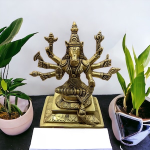 May include: A gold-colored brass statue of a Hindu deity with multiple arms, sitting on a square base. The deity has a boar-like face and is adorned with intricate details.