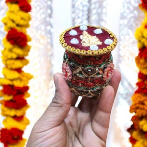May include: A small, round, red and gold decorative pot with a lid. The lid is decorated with white beads and a gold leaf design. The pot is decorated with intricate patterns and beads.