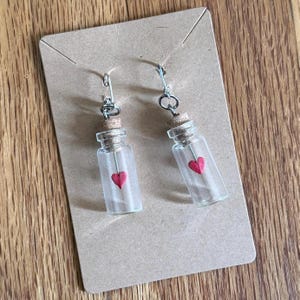 May include: A pair of glass bottle earrings with cork stoppers. Each clear bottle contains a small red heart. The earrings hang from silver-colored hooks and are displayed on a brown card.