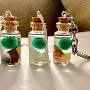 May include: Three small glass bottles with cork stoppers and silver chains. Each bottle contains a green moss ball, clear liquid, and colorful stones. The bottles are attached to key rings.