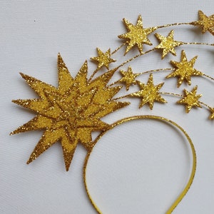 May include: A gold headband with a large glitter star and smaller glitter stars attached to it. The headband is made of a thin, flexible material.