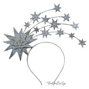May include: Silver glitter star headband with a large star in the center and smaller stars trailing behind it. The headband is white and has the text "Dekor Livsy" printed on it.