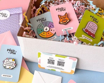 May include: A box filled with colourful shredded paper and enamel pins. The pins feature various designs, including a fox with the text "Be Kind to All Kinds", a tombstone with the text "Here Lies The Last Fuckboy Who Tried", a spinning wheel with the text "Petit + Pin", and a hand with the text "Loved".
