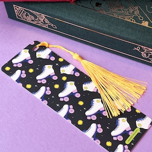 Könnte beinhalten: Ein schwarz-weißes Lesezeichen mit einem sich wiederholenden Muster aus weißen Rollschuhen mit gelben Smileys auf schwarzem Hintergrund. Das Lesezeichen hat einen gelben Quasten.