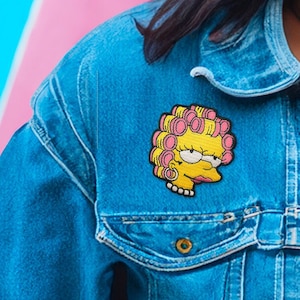 Peut inclure: Une veste en jean bleue avec un patch de personnage de dessin animé jaune et rose sur le côté gauche de la poitrine. Le personnage a des rouleaux de cheveux roses et un visage jaune.