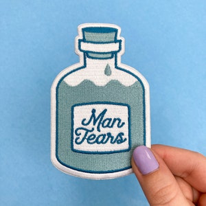 May include: A blue and white embroidered patch of a bottle with a label that reads "Man Tears".