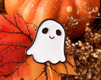 May include: A white enamel pin shaped like a ghost with black eyes and a smiling mouth. The pin is on a background of orange and brown fall leaves.
