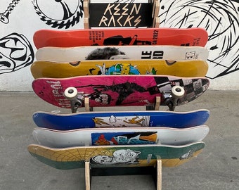 Könnte beinhalten: Ein Skateboard-Ständer aus Holz mit sieben Skateboards. Die Skateboards haben verschiedene farbenfrohe Designs und Grafiken. Der Ständer ist mit "Keen Racks" beschriftet.