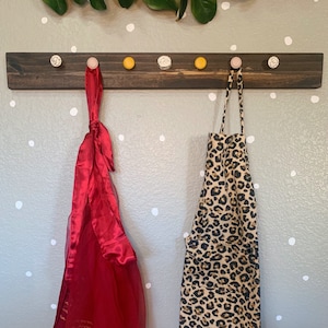 May include: A wooden wall-mounted rack with colorful knobs, holding a red and gold scarf and a leopard print apron.