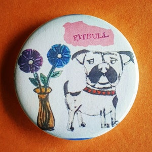May include: A white button with a cartoon illustration of a dog with the word "Pitbull" written on a pink banner above the dog. The dog is black and white with a red collar. The dog is standing next to a vase with three flowers, two purple and one blue.