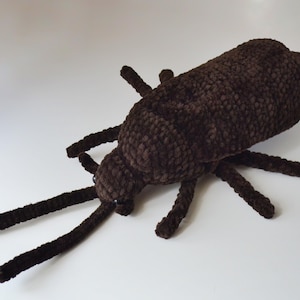May include: A dark brown crochet beetle plush toy featuring a textured yarn surface, six segmented legs, and two long, thin antennae extending from the head. The insect-shaped stuffed animal has small black bead eyes and a rounded body structure.