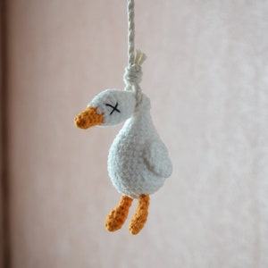 Puede incluir: Un ganso blanco de crochet con patas naranjas colgando de una cuerda blanca. El ganso tiene una X negra para los ojos.