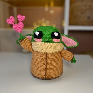 Pu&ograve; includere: Bambola all'uncinetto fatta a mano che assomiglia a un personaggio di una popolare serie di fantascienza. La bambola &egrave; principalmente marrone, verde e beige, con dettagli rosa. Tiene in mano un piccolo mazzo di cuori rosa.