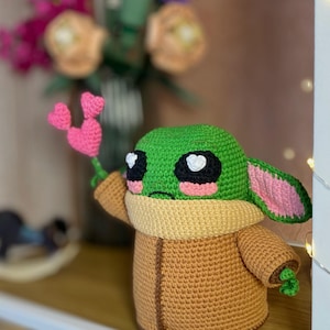 Pu&ograve; includere: Una figura all'uncinetto di un personaggio con la testa verde, grandi occhi neri e guance rosa, che tiene in mano un fiore a forma di cuore rosa. La figura indossa un cappotto marrone e un colletto color crema. Lo sfondo include fiori.