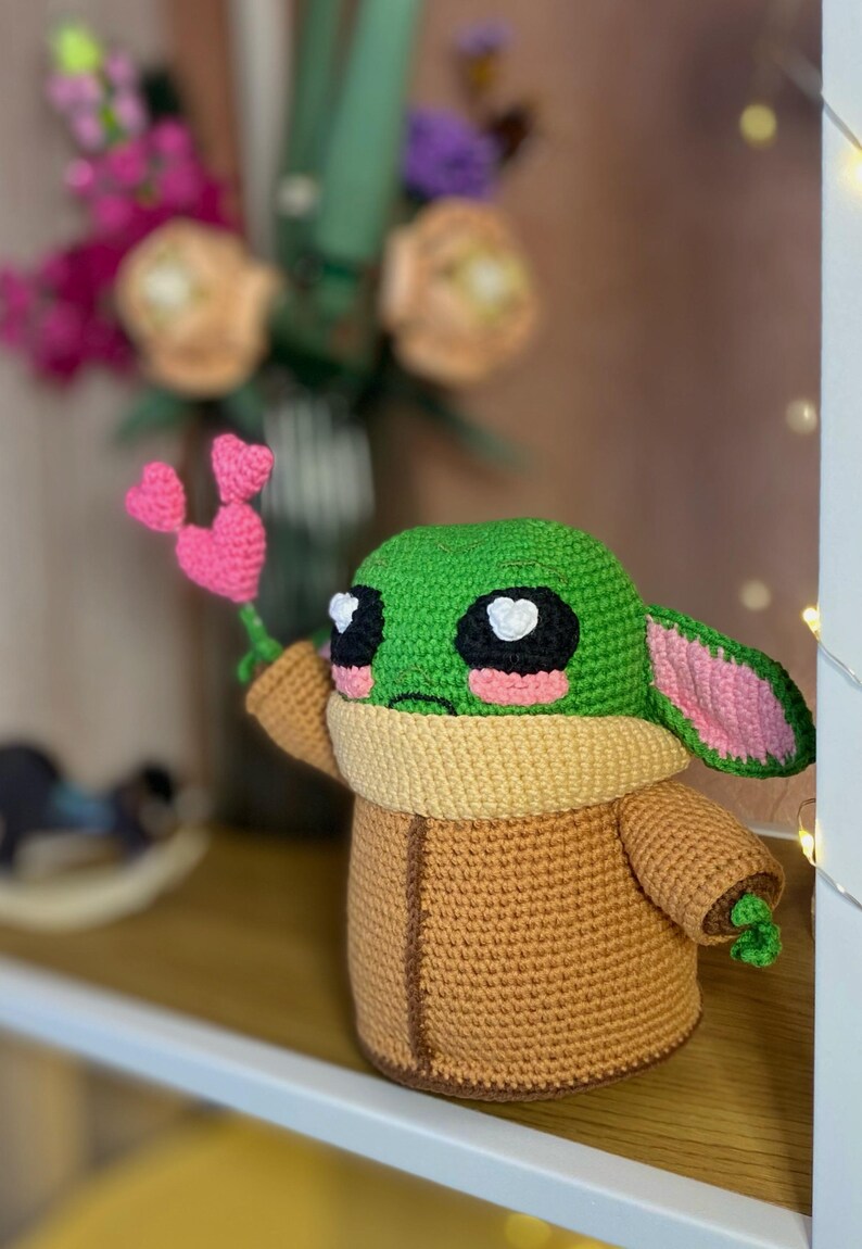 Pu&ograve; includere: Una figura all'uncinetto di un personaggio con la testa verde, grandi occhi neri e guance rosa, che tiene in mano un fiore a forma di cuore rosa. La figura indossa un cappotto marrone e un colletto color crema. Lo sfondo include fiori.
