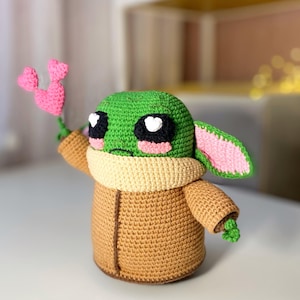 Pu&ograve; includere: Bambola all'uncinetto fatta a mano a somiglianza di un personaggio popolare, con testa verde, grandi occhi neri con riflessi bianchi a forma di cuore e una veste color cammello. La bambola tiene in mano un fiore rosa a forma di cuore. La bambola &egrave; alta circa 15 cm.
