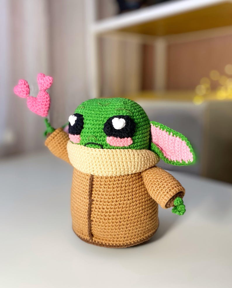 Pu&ograve; includere: Bambola all'uncinetto fatta a mano a somiglianza di un personaggio popolare, con testa verde, grandi occhi neri con riflessi bianchi a forma di cuore e una veste color cammello. La bambola tiene in mano un fiore rosa a forma di cuore. La bambola &egrave; alta circa 15 cm.