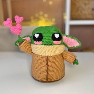 Pu&ograve; includere: Bambola all'uncinetto fatta a mano, raffigurante un personaggio popolare, che tiene in mano un mazzo di cuori rosa. La bambola ha una testa verde, grandi occhi neri e un cappotto marrone. Le orecchie sono foderate di rosa e la bambola ha un scaldacollo marrone.