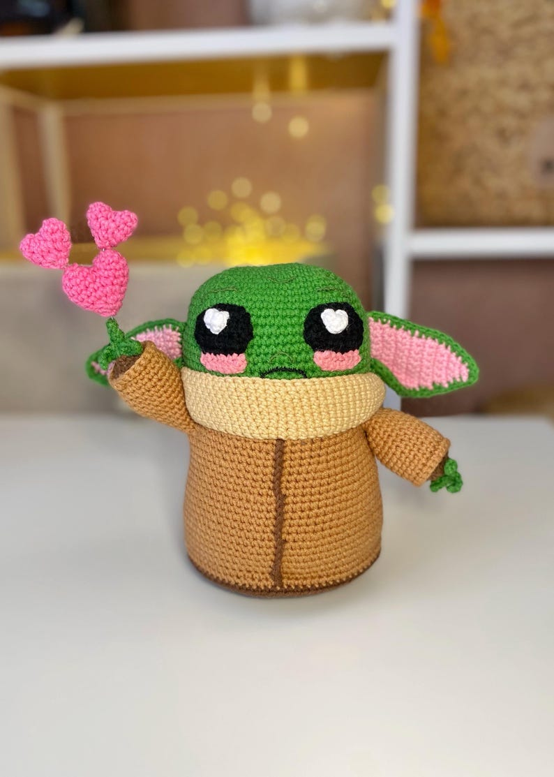 Pu&ograve; includere: Bambola all'uncinetto fatta a mano, raffigurante un personaggio popolare, che tiene in mano un mazzo di cuori rosa. La bambola ha una testa verde, grandi occhi neri e un cappotto marrone. Le orecchie sono foderate di rosa e la bambola ha un scaldacollo marrone.