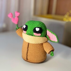 Pu&ograve; includere: Una figura all'uncinetto di un personaggio con la testa verde, grandi occhi neri e bianchi e un cappotto marrone. La figura tiene in mano un fiore rosa a forma di cuore. La figura ha un colletto marrone e orecchie foderate di rosa. &Egrave; un personaggio popolare.