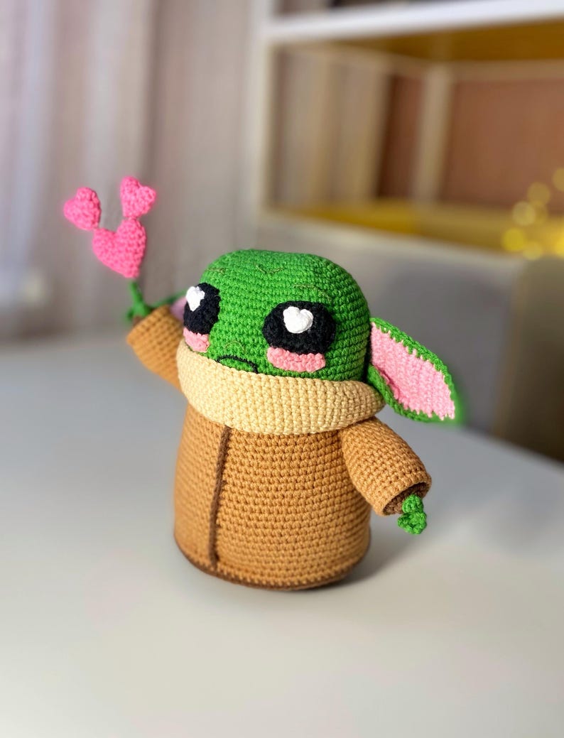 Pu&ograve; includere: Una figura all'uncinetto di un personaggio con la testa verde, grandi occhi neri e bianchi e un cappotto marrone. La figura tiene in mano un fiore rosa a forma di cuore. La figura ha un colletto marrone e orecchie foderate di rosa. &Egrave; un personaggio popolare.