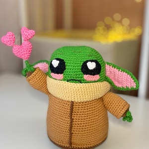 Pu&ograve; includere: Bambola all'uncinetto fatta a mano che assomiglia a un personaggio popolare, con testa verde, grandi occhi neri e guance rosa. La bambola indossa un cappotto marrone e tiene in mano un gruppo di cuori rosa all'uncinetto. Le orecchie sono rosa e verdi.