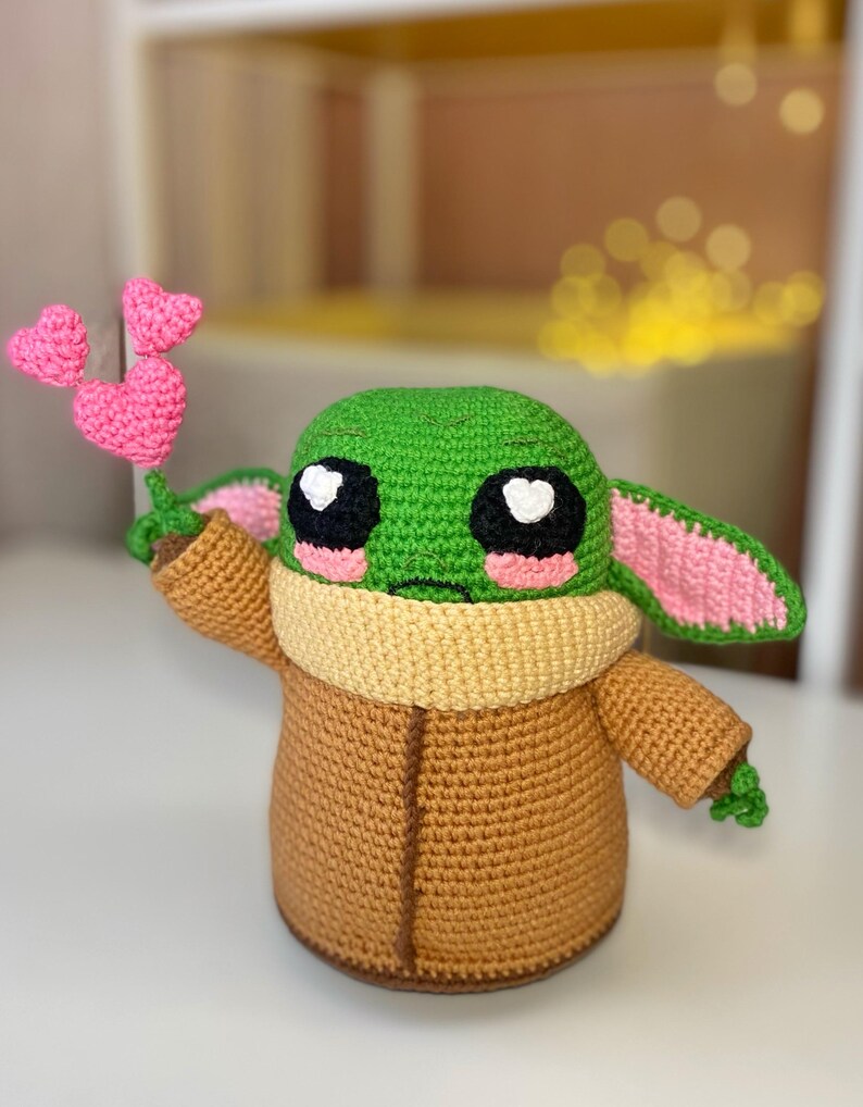 Pu&ograve; includere: Bambola all'uncinetto fatta a mano che assomiglia a un personaggio popolare, con testa verde, grandi occhi neri e guance rosa. La bambola indossa un cappotto marrone e tiene in mano un gruppo di cuori rosa all'uncinetto. Le orecchie sono rosa e verdi.