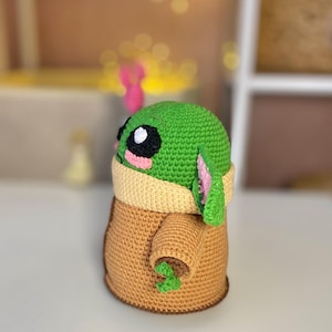 Pu&ograve; includere: Una figura all'uncinetto fatta a mano di un personaggio con una testa grande, grandi occhi neri e pelle verde. La figura indossa una veste marrone con un colletto color crema. La figura ha grandi orecchie e piccole mani verdi. La figura &egrave; alta circa 15 cm.