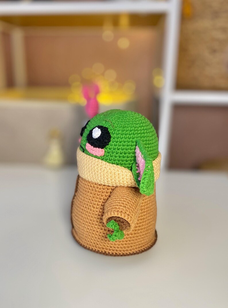 Pu&ograve; includere: Una figura all'uncinetto fatta a mano di un personaggio con una testa grande, grandi occhi neri e pelle verde. La figura indossa una veste marrone con un colletto color crema. La figura ha grandi orecchie e piccole mani verdi. La figura &egrave; alta circa 15 cm.
