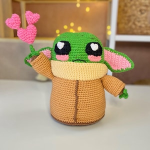 Pu&ograve; includere: Una figura all'uncinetto di un personaggio con la testa verde, grandi occhi neri e guance rosa, che tiene in mano tre palloncini rosa a forma di cuore. La figura indossa un cappotto marrone e un colletto color crema.