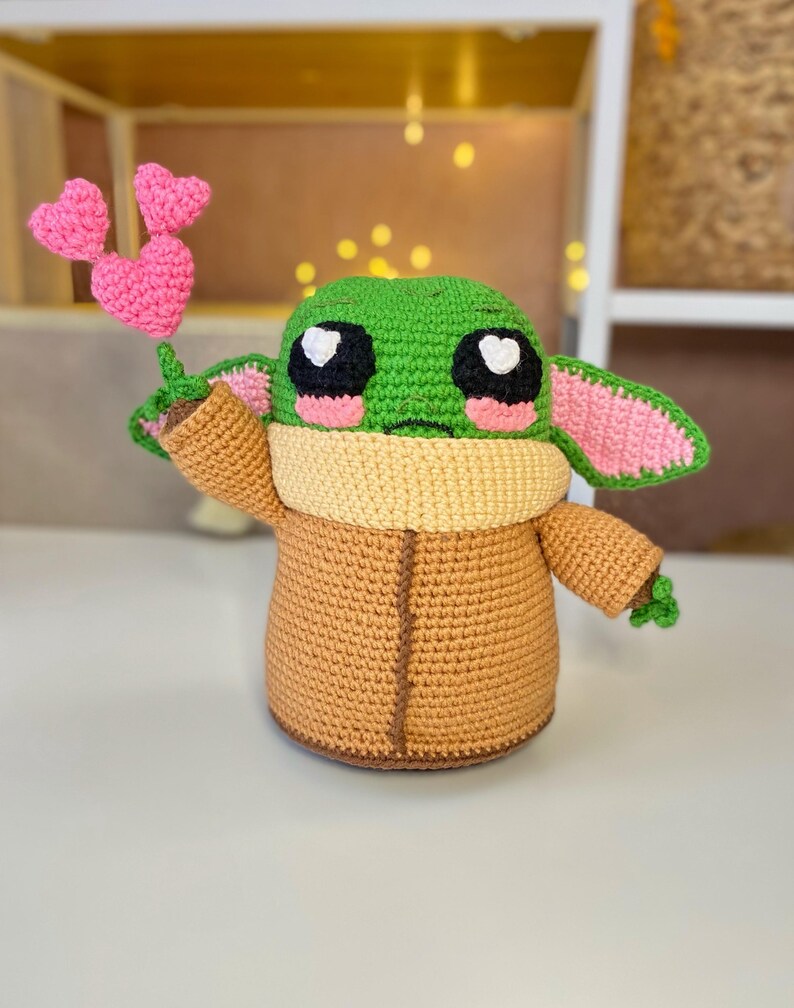 Pu&ograve; includere: Una figura all'uncinetto di un personaggio con la testa verde, grandi occhi neri e guance rosa, che tiene in mano tre palloncini rosa a forma di cuore. La figura indossa un cappotto marrone e un colletto color crema.