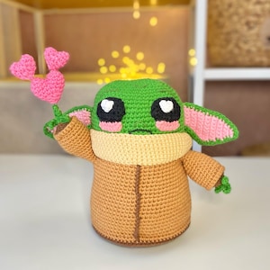 Pu&ograve; includere: Bambola all'uncinetto fatta a mano che assomiglia a un personaggio popolare, con una testa verde, grandi occhi bianchi e neri e dettagli rosa. La bambola indossa un cappotto marrone e tiene in mano un piccolo bouquet di cuori rosa all'uncinetto.
