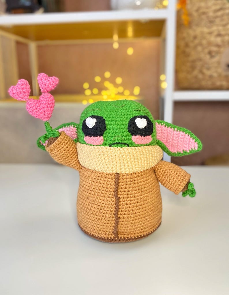 Pu&ograve; includere: Bambola all'uncinetto fatta a mano che assomiglia a un personaggio popolare, con una testa verde, grandi occhi bianchi e neri e dettagli rosa. La bambola indossa un cappotto marrone e tiene in mano un piccolo bouquet di cuori rosa all'uncinetto.