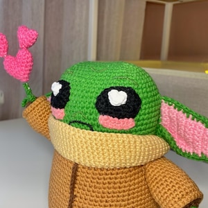 Pu&ograve; includere: Una figura all'uncinetto di un personaggio dalla pelle verde con grandi occhi neri, guance rosa e grandi orecchie. La figura indossa un cappotto marrone e un colletto giallo. Tiene in mano un fiore a forma di cuore rosa all'uncinetto.
