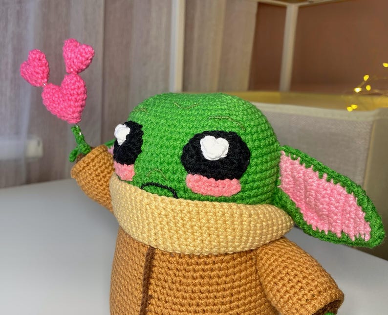Pu&ograve; includere: Una figura all'uncinetto di un personaggio dalla pelle verde con grandi occhi neri, guance rosa e grandi orecchie. La figura indossa un cappotto marrone e un colletto giallo. Tiene in mano un fiore a forma di cuore rosa all'uncinetto.
