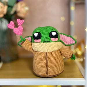 Pu&ograve; includere: Una figura all'uncinetto fatta a mano di un personaggio con grandi occhi e pelle verde, che indossa una veste marrone e tiene oggetti a forma di cuore rosa. La figura &egrave; esposta su uno scaffale con uno sfondo sfocato.