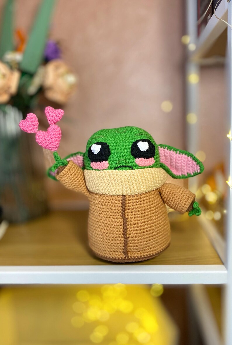 Pu&ograve; includere: Una figura all'uncinetto fatta a mano di un personaggio con grandi occhi e pelle verde, che indossa una veste marrone e tiene oggetti a forma di cuore rosa. La figura &egrave; esposta su uno scaffale con uno sfondo sfocato.