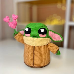 Pu&ograve; includere: Figura all'uncinetto di un personaggio con grandi occhi neri, testa verde e corpo marrone. La figura tiene in mano un gruppo di tre cuori rosa all'uncinetto. La figura ha grandi orecchie con interni rosa e un colletto beige.