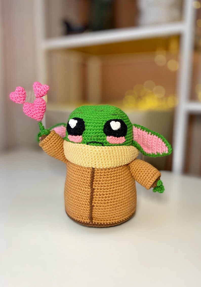 Pu&ograve; includere: Figura all'uncinetto di un personaggio con grandi occhi neri, testa verde e corpo marrone. La figura tiene in mano un gruppo di tre cuori rosa all'uncinetto. La figura ha grandi orecchie con interni rosa e un colletto beige.