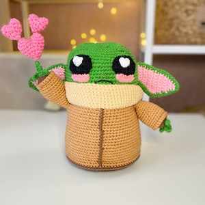 Pu&ograve; includere: Bambola all'uncinetto di un personaggio con grandi occhi neri e bianchi, testa verde e veste marrone. La bambola tiene in mano tre cuori rosa all'uncinetto. La bambola ha guance e orecchie rosa e un colletto marrone.