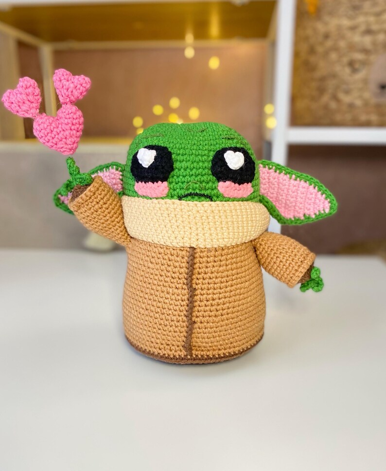 Pu&ograve; includere: Bambola all'uncinetto di un personaggio con grandi occhi neri e bianchi, testa verde e veste marrone. La bambola tiene in mano tre cuori rosa all'uncinetto. La bambola ha guance e orecchie rosa e un colletto marrone.