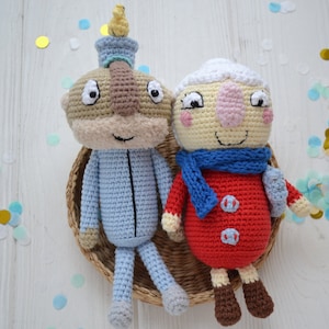May include: Two crocheted plush toys, one blue and one red, sitting in a wicker basket. The blue toy has a brown face and a yellow bird on its head. The red toy has a white face and a blue scarf. The toys are surrounded by confetti.