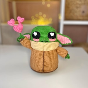Pu&ograve; includere: Bambola all'uncinetto di un personaggio con grandi occhi neri, pelle verde e grandi orecchie. La bambola tiene in mano un fiore rosa a forma di cuore. La bambola indossa un cappotto marrone e un colletto beige. La bambola &egrave; alta circa 20 cm.