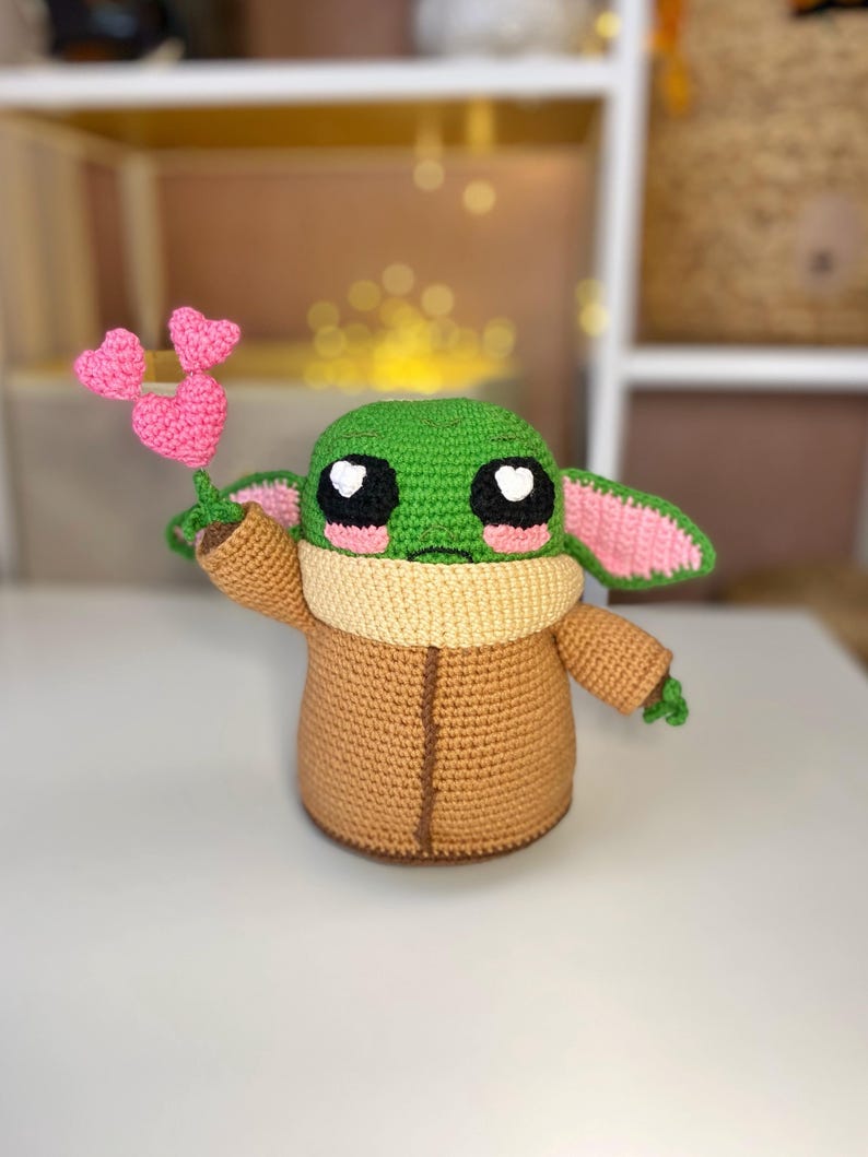 Pu&ograve; includere: Bambola all'uncinetto di un personaggio con grandi occhi neri, pelle verde e grandi orecchie. La bambola tiene in mano un fiore rosa a forma di cuore. La bambola indossa un cappotto marrone e un colletto beige. La bambola &egrave; alta circa 20 cm.