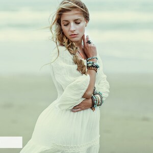 May include: A woman with long blonde hair wearing a white dress and multiple bracelets on her wrist. She is standing on a beach with the ocean in the background.