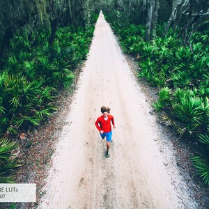 May include: A person in a red shirt and blue shorts runs down a dirt path lined with green foliage.