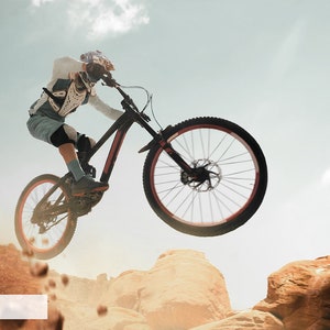 May include: A person wearing protective gear rides a black mountain bike over a rocky terrain. The bike is in mid-air, with the front wheel off the ground. The rider is wearing a helmet, goggles, and a long-sleeved shirt. The background is a blue sky with white clouds.
